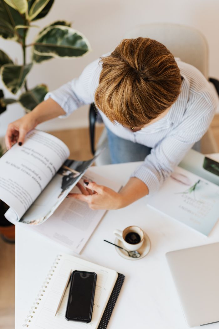 From above anonymous female in jeans and light blue shirt working at desk with gadgets and papers while turning over magazine and having cup of hot americano